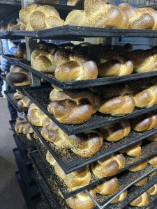 Trays of freshly baked sesame challah loaves stacked on black bakery racks in Bnei Brak.