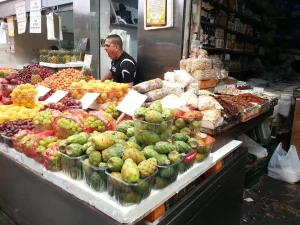 A produce stall piled with grapes, sabras, plums and dried fruit at Mahane Yehuda market in Jerusalem, a stallholder calling out behind it.