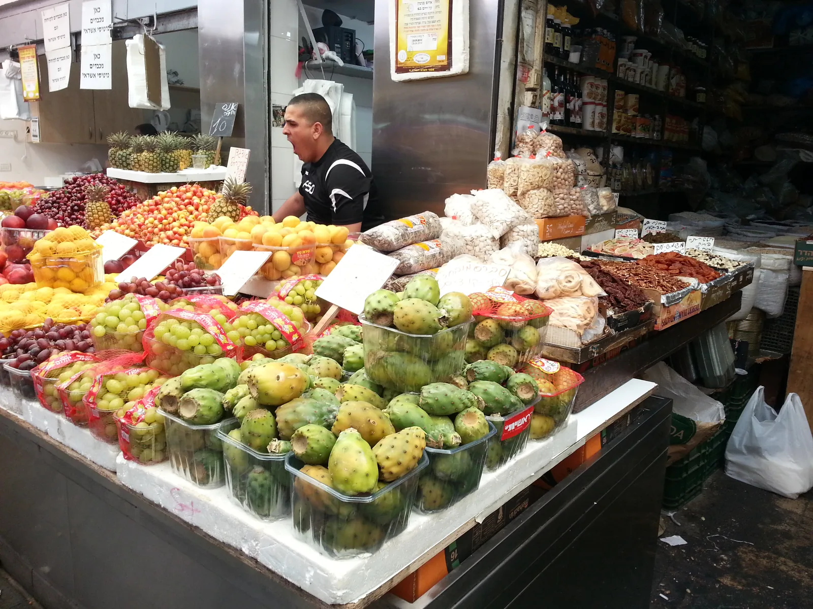A produce stall piled with grapes, sabras, plums and dried fruit at Mahane Yehuda market in Jerusalem, a stallholder calling out behind it.