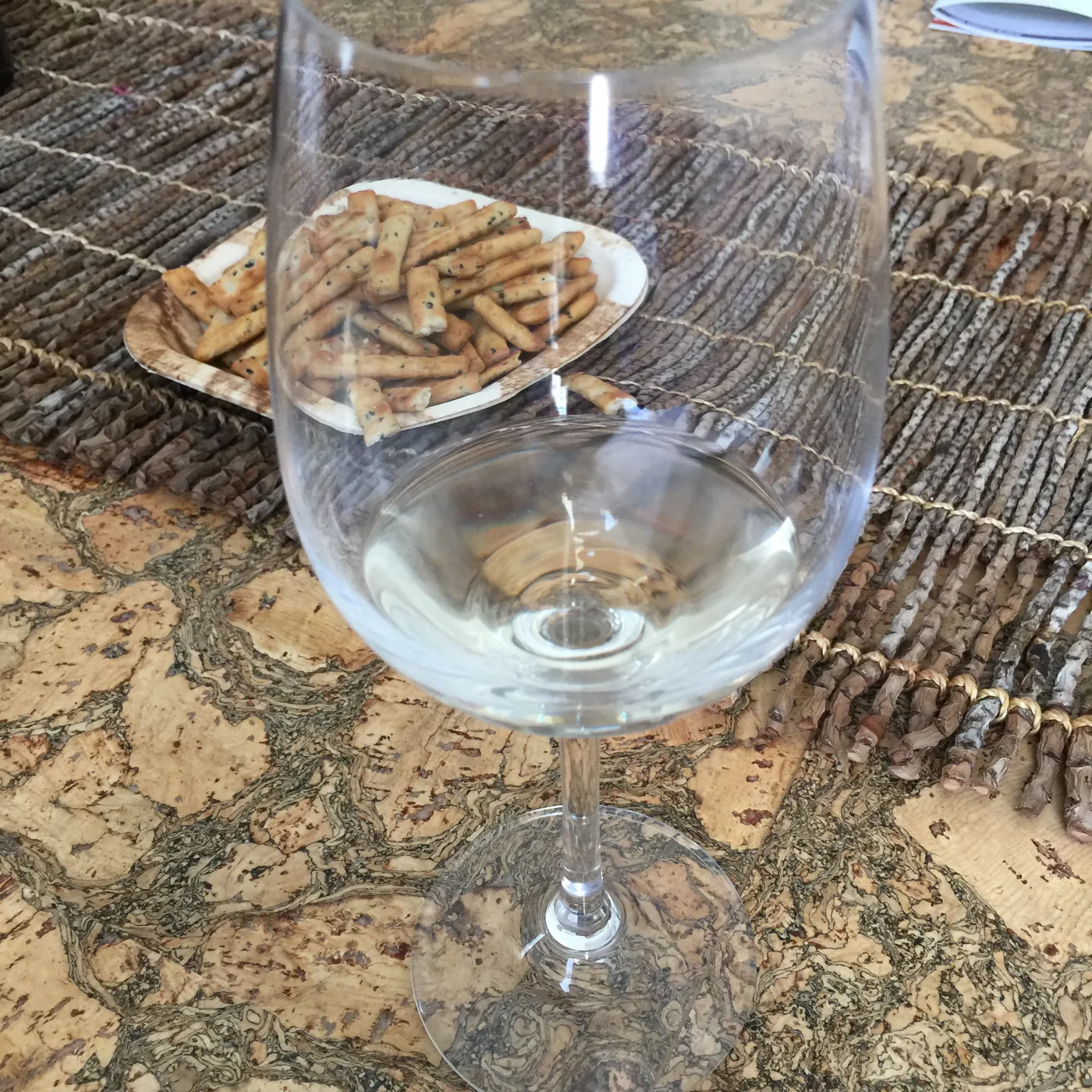 A filled glass of white wine on a cork placemat beside a small dish of sesame snacks