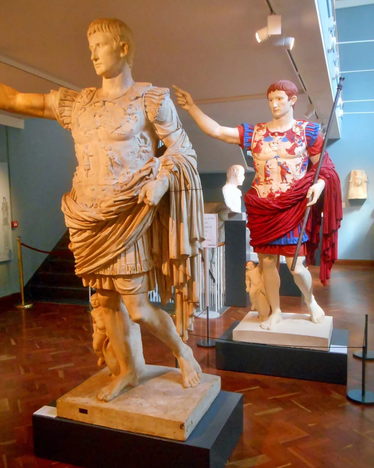 A white statue of the Roman emperor Augustus in the foreground, with a brightly painted copy of the same statue in red and blue behind it, in a museum gallery