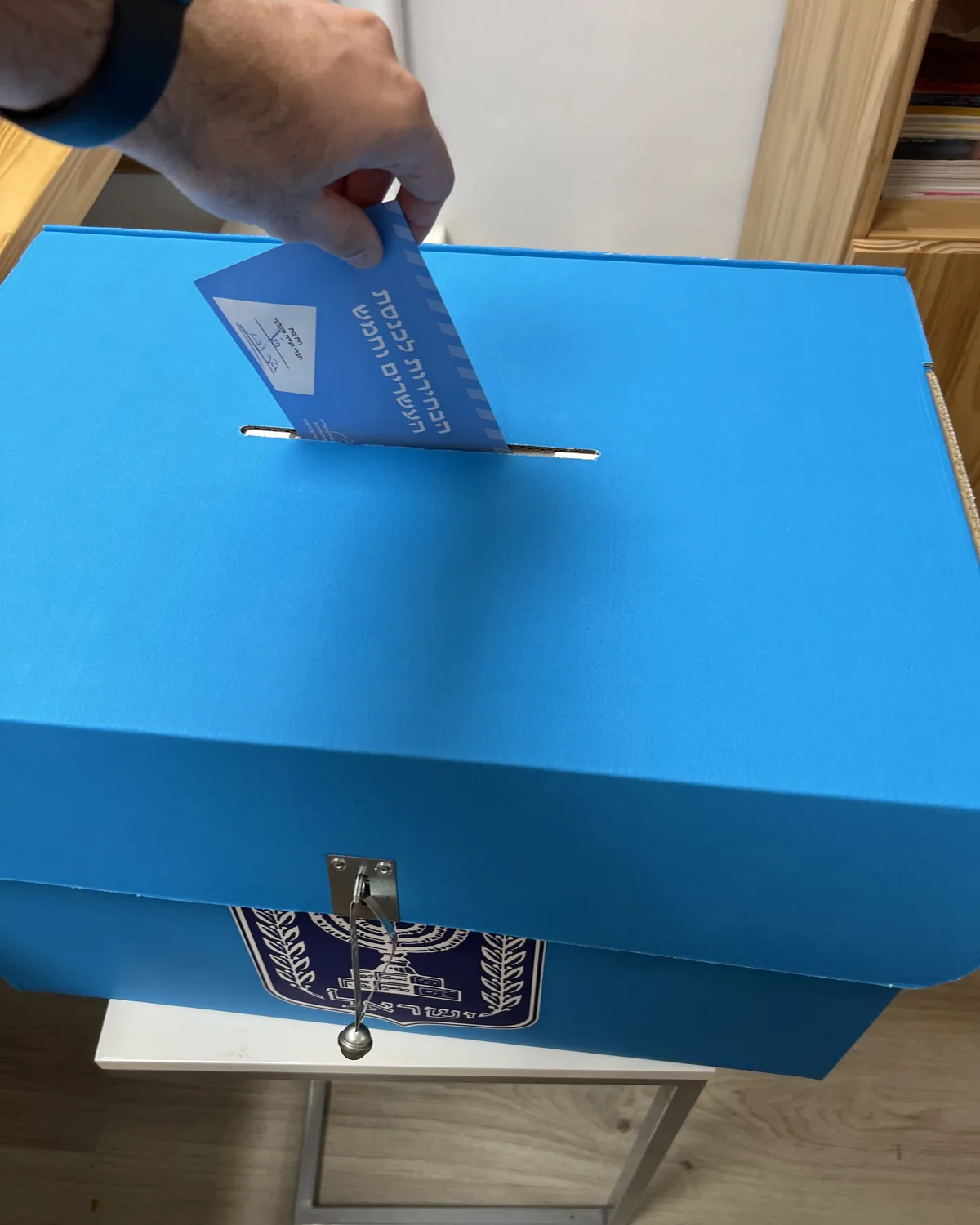 A hand posting a blue election envelope into a blue ballot box marked with the State of Israel emblem.