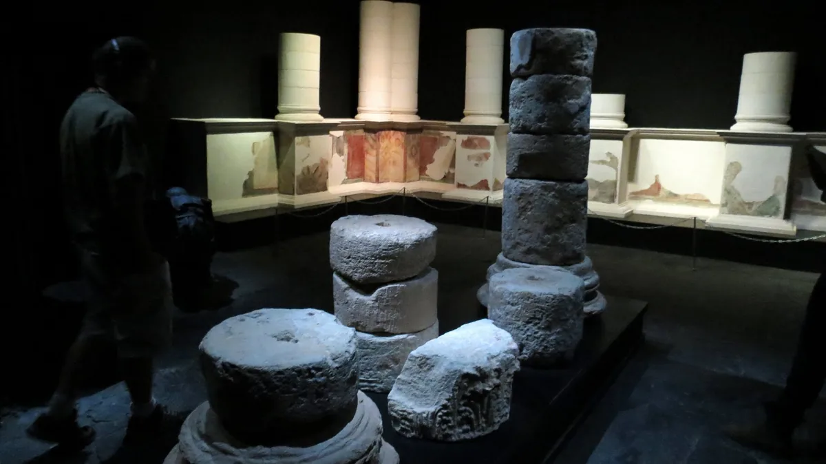 Painted wall panels in red, yellow and black, with stacked stone column sections from Masada, in a dimly lit museum gallery